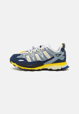 Zapatilla de running con parte superior de malla gris, acentos en azul marino y amarillo, suela texturizada, detalles reflectantes y un cierre de cordones seguro.