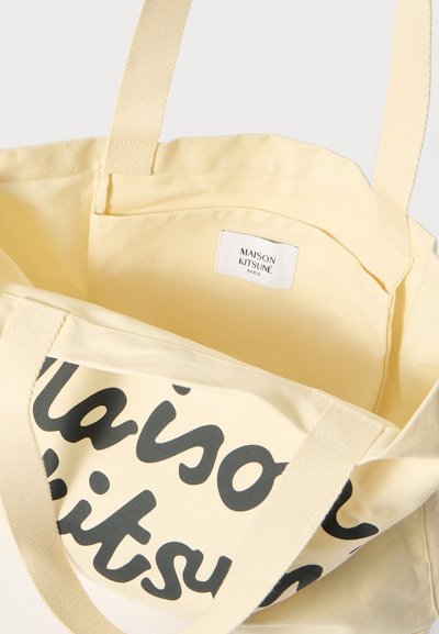 Žlutá bavlněná taška přes rameno s černým textem "maison kitsuné" uvnitř. Obsahuje textilní ucha a malou vnitřní etiketu. Měkká struktura.