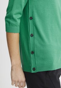 Zeleno pleteno oblačilo s krojaškim ovratnikom, rokavi do tri četrtine in črnimi gumbi ob levi strani. Gladka tekstura.