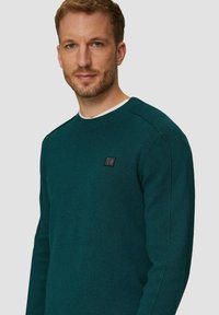 Teal gebreide trui met een ronde halslijn, voorzien van een klein zwart logo op de borst. Zachte textuur, lange mouwen en geribbelde manchetten.