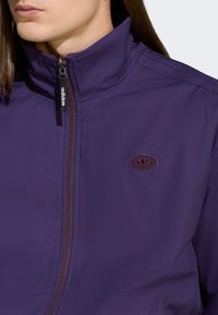 Violetti vetoketjullinen takki, joka on valmistettu kevyestä kankaasta. Ominaisuuksia ovat korkea kaulus, ruskea vetoketju ja kirjailtu logo rinnassa.