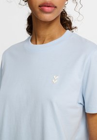 Ljusblå bomulls t-shirt med rund halsringning, med en liten krämfärgad chevronlogotyp på vänster bröstområde.