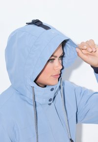 Ljust blå väderbeständig jacka med en stor huva, justerbara remmar, marinblå detaljer och vita dragsnören. Slät tyg med en figursydd form.