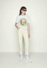 Pessoa vestindo uma t-shirt gráfica branca "Casablanca", calças amarelas claras, sapatos brancos e óculos de sol escuros, de pé contra um fundo simples.