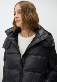Doudoune noire avec un col montant, fermeture éclair et accents de boutons. Texture lisse et brillante avec un design matelassé et poches avant.