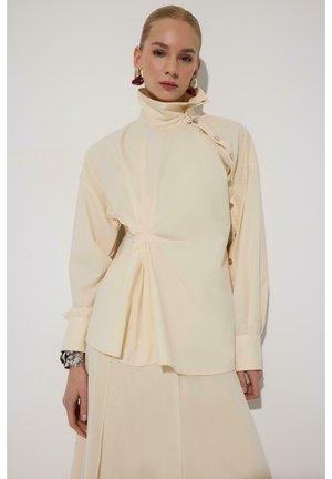 Femeie purtând o bluză crem cu guler înalt, nasturi laterali și pliseuri asimetrice, asortată cu o fustă similară și cercei statement.