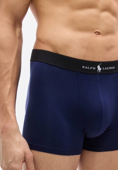 Ανδρικός κορμός που φοράει μπλε σκουρόχρωμα μποξεράκια Ralph Lauren με μαύρη μέση και λογότυπο, παρουσιάζοντας γυμνασμένη κοιλιά και το αριστερό χέρι δίπλα στο σώμα.