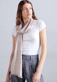Beżowy szalik wykonany z miękkiego materiału, z delikatnym wzorem. Szalik jest zarzucony na szyję, z frędzlami na kraw�ędziach.