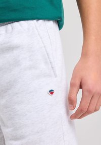 Pantaloni della tuta grigi chiari con una texture morbida, caratterizzati da un piccolo logo triangolare ricamato in blu, rosso e bianco sul lato sinistro.
