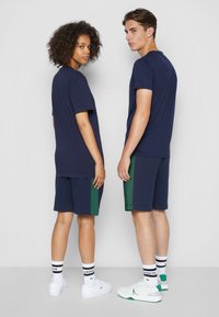 Marine blousons gecombineerd met marine korte broeken met groene zijkanten. Beide dragen witte sneakers en gestreepte sokken. Van een katoenmix materiaal.