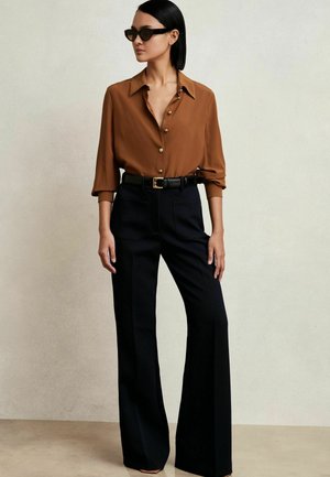 Camicia marrone con bottoni e colletto, abbinata a pantaloni neri a vita alta. Gli accessori includono una cintura nera e occhiali da sole.