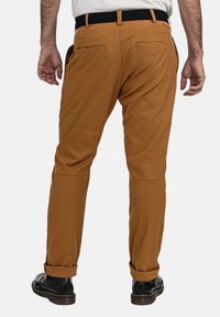 Braune, leichte Chinos mit geradem Bein, zwei Gesäßtaschen und hochgekrempelten Säumen, kombiniert mit schwarzen Schuhen mit gelben Nähten.