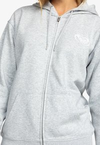 Femme portant un sweat à capuche gris clair zippé avec des poches avant et un petit logo blanc sur le côté gauche de la poitrine.