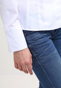 Valkoinen pitkähihainen paita napitettuine hihoineen, yhdistettynä sinisiin farkkuihin. Käsi lepää farkuilla, mikä osoittaa sileää kangasta ja hyvin istuvaa leikkausta.