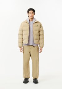 Chaqueta acolchada beige con textura de pana, combinada con un polo gris claro y pantalones beige, llevada con zapatillas negras estampadas.
