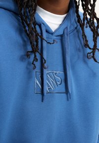 Personne portant un sweat à capuche Vans bleu avec cordons par-dessus une chemise blanche, montrant des cheveux tressés tombant devant.