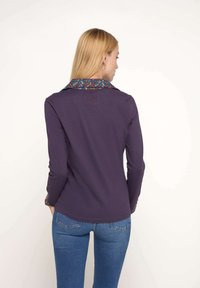 Chemise à manches longues violette avec un col à motif bleu, orange et marron. Elle présente un ourlet arrondi et des détails brodés au dos.