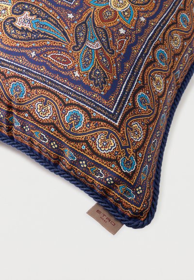 Coin de coussin décoratif avec un motif cachemire complexe en tons de bleu, marron et or, bordé d'une tresse marine et d'une étiquette beige portant l'inscription "ETRO HOME".