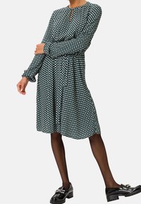 Temně zelené šaty s bílým geometrickým vzorem, dlouhými rukávy, detaily ve tvaru klíčové dírky, nařaseným pasem, v kombinaci s černými lakovanými mokasíny a punčochami.