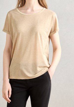 Frau trägt ein beigefarbenes kurzärmeliges T-Shirt, das leicht in schwarze Hose gesteckt ist, steht mit einer Hand in der Tasche vor einem schlichten hellen Hintergrund.