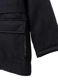 Primer plano de la manga y el bolsillo con tapa de una chaqueta negra, mostrando costuras detalladas y un atisbo del forro interior a cuadros.