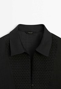 Chemise noire à col avec un panneau avant tissé texturé et un bouton visible unique en bas de la patte de boutonnage.
