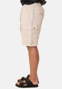 Pantaloni cargo beige in lino con tasche laterali e tasche posteriori a patta. Vita elastica con cordoncino. Indossati con sandali neri.