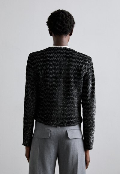 Black shimmer jacket with a chevron pattern, textured fabric, round neckline, and long sleeves, paired with gray high-waisted trousers.