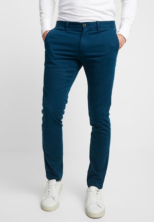 Mann trägt dunkelblaue schlanke Hose, weiße Sneaker und ein weißes Langarmshirt, die Hände in den Taschen.