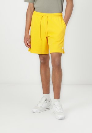 Mann trägt leuchtend gelbe Shorts mit Kordelzug, weiße Socken und weiße Turnschuhe, steht vor einem schlichten weißen Hintergrund.