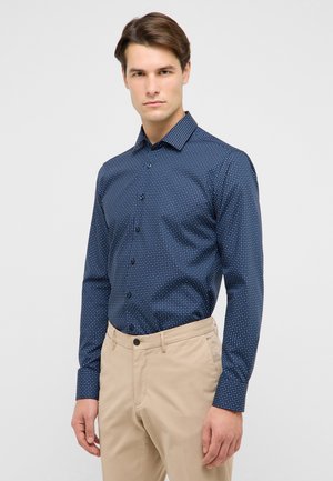 Marineblaue Hemdbluse mit weißen Polka-Dots, langen Ärmeln und klassischem Kragen, kombiniert mit hellbeigen Hosen. Glatte Stoffstruktur.