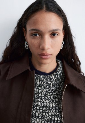 Mladi žena s dugom tamnom kosom, koja nosi naušnice od srebrne lanca, crno-bijeli pleteni džemper i tamnobraon jaknu na bijeloj pozadini.