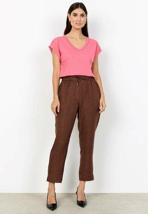 Vrouw draagt een roze top met korte mouwen en bruine hoge taillebroek, staand tegen een effen witte muur met zwarte hakken.