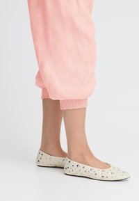 Ballerines en cuir blanc cassé, ornées de clous argentés et à bout arrondi. Associées à un pantalon évasé rose à effet élastique.