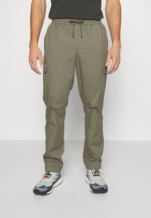 Homme portant un pantalon cargo vert olive avec taille à cordon et poches latérales, associé à des baskets grises et blanches.