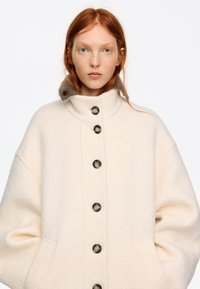 Mujer joven de piel clara y cabello rojo lleva un abrigo de lana crema oversize con grandes botones y cuello alto, posando frente a un fondo blanco.