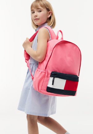 Ragazza giovane in vestito a righe che porta uno zaino grande rosa con tasca anteriore rossa, bianca e blu navy, in piedi davanti a uno sfondo semplice.