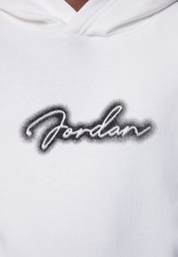 Felpa bianca con cappuccio caratterizzata da un logo in testo nero spruzzato, "Jordan", realizzata in un morbido tessuto texturizzato e con una tasca a marsupio frontale.
