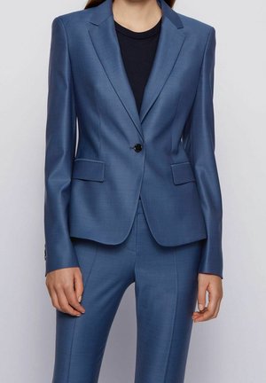Kvinde iført en skræddersyet blå blazer med matchende bukser og en sort top mod en ensfarvet baggrund.