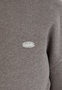 Bruine sweatshirt met een zachte textuur, met een zilveren ovale logo plaat die "Smulblox" zegt, bevestigd met metalen klinknagels.