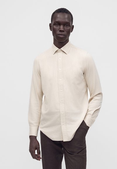 Homme à la peau foncée portant une chemise beige clair boutonnée et un pantalon foncé, debout devant un fond gris clair uni.
