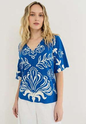 Kvinde med bølget blondt hår iført en blå V-hals bluse med hvide blomsterprint og hvide bukser, stående mod en ensfarvet baggrund.