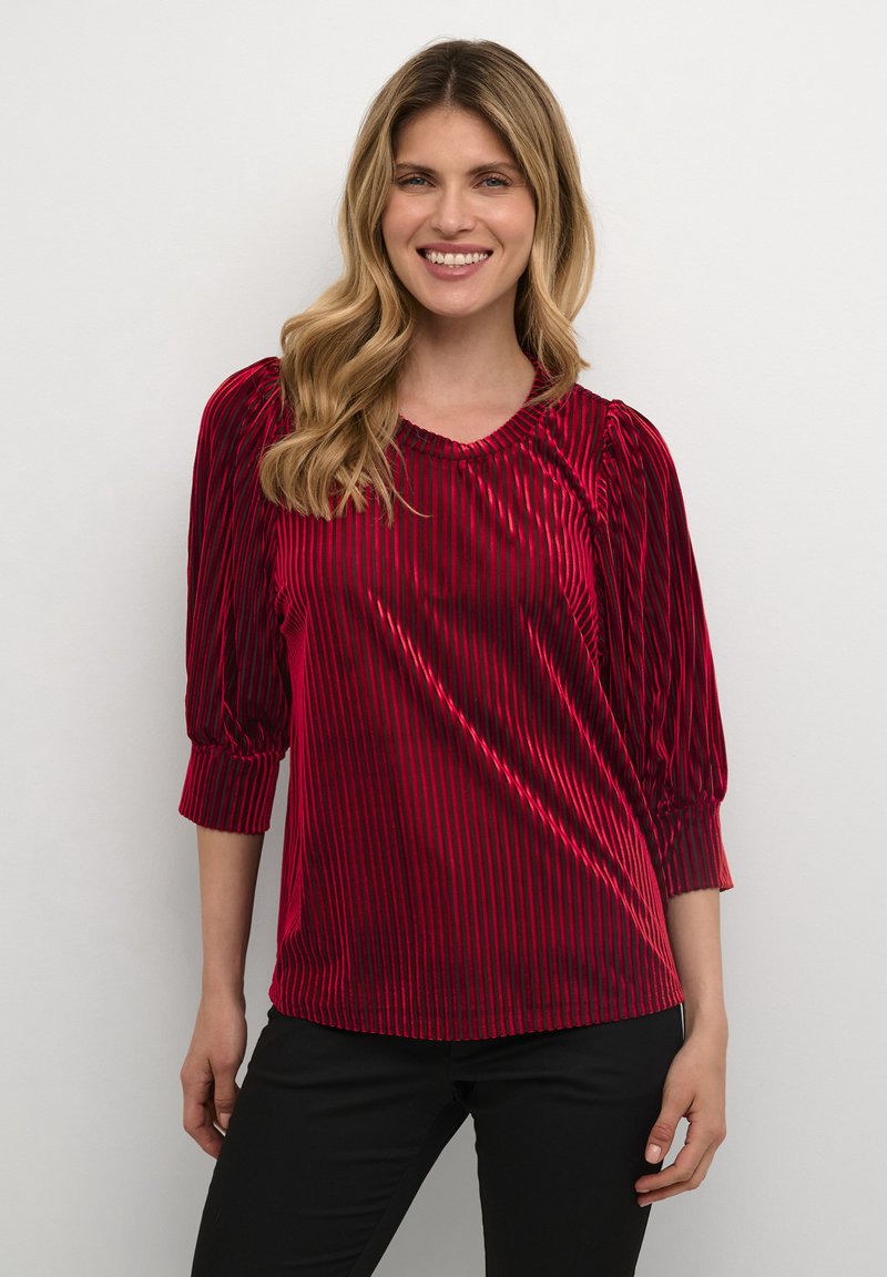 Blusa in velluto rosso con righe verticali, scollatura rotonda e maniche a sbuffo a tre quarti. Il tessuto ha un aspetto morbido e strutturato.