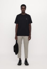Tricou din bumbac negru cu un mic logo multicolor pe piept, asortat cu pantaloni gri deschis și pantofi negri.