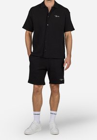 Set van zwart shirt met korte mouwen en shorts, met een getextureerd ruitpatroon, knoopsluiting en geborduurd logo. Gecombineerd met witte sneakers.