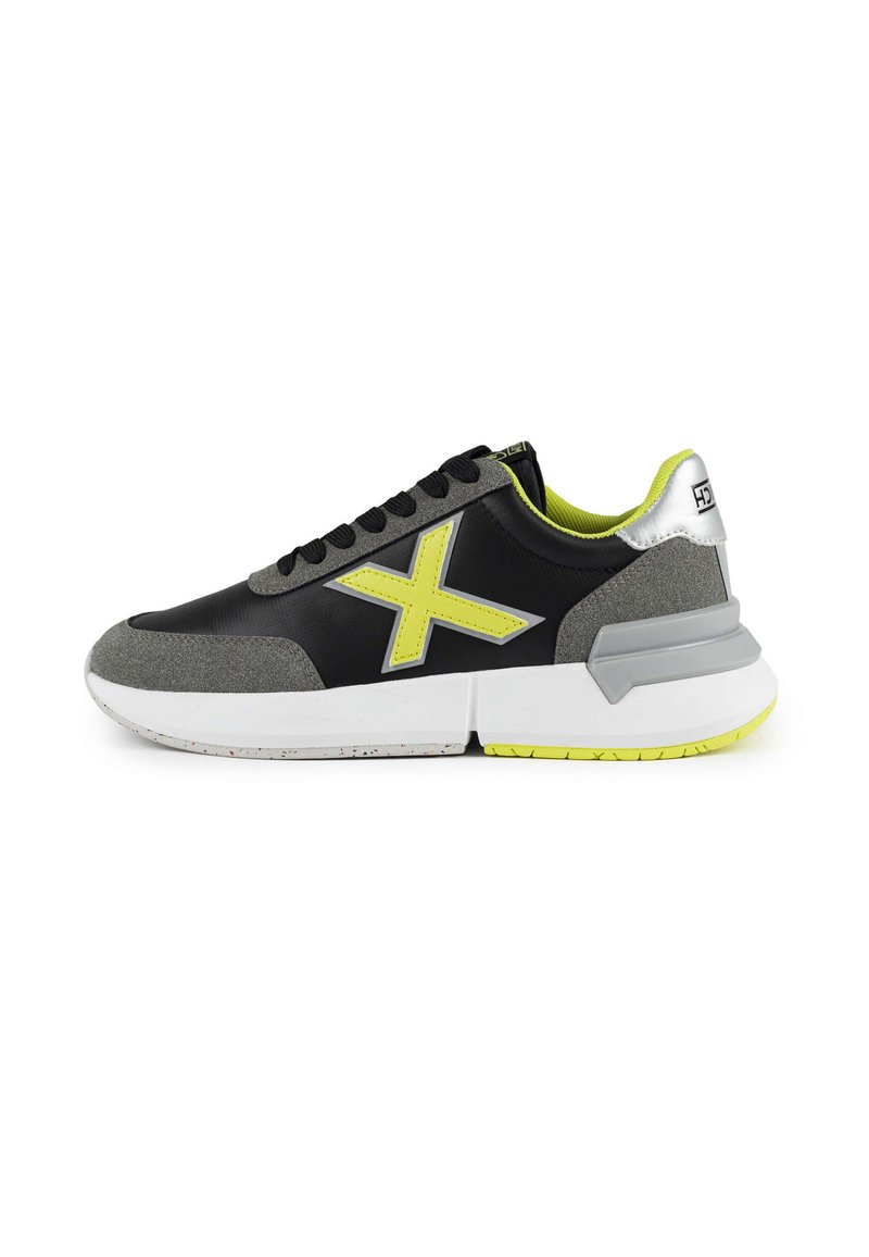 Chaussure de sport noire et grise avec un logo jaune fluo, des accents en daim texturé et une semelle intermédiaire blanche. Lacets et col rembourré inclus.