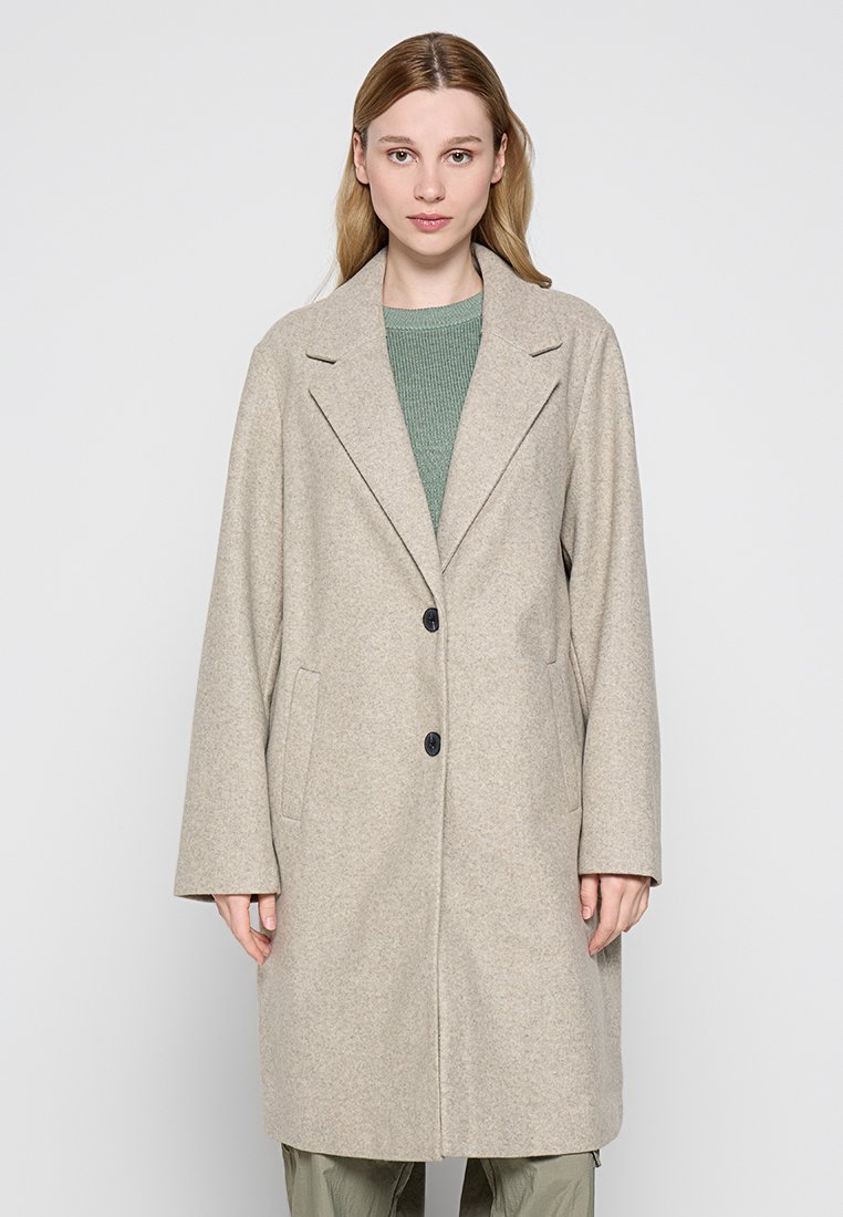 Mujer vistiendo un abrigo largo de lana beige sobre un suéter verde y pantalones oliva, de pie contra un fondo claro y liso.