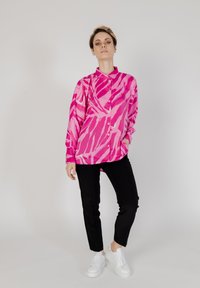 Camisa de manga larga en rosa brillante con un patrón de cebra abstracto, con un cuello abotonado. Combinada con pantalones negros y zapatillas blancas.