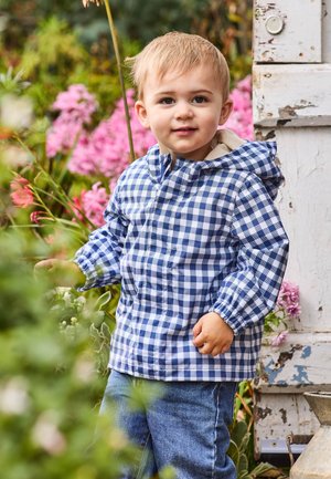 Tout-petit aux cheveux blonds portant une veste à carreaux bleue et blanche et un jean, se tient près de fleurs roses et d'une porte blanche usée à l'extérieur.