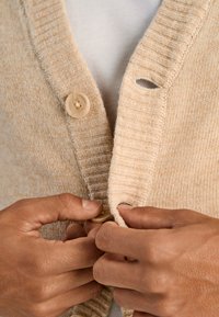 Beige stickad kofta med ribbad krage och knappstängning. Har ett litet hål nära nackområdet, händer knäpper knappen.